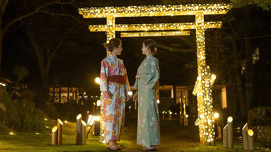 浴衣を着た2人の女性が、光で装飾された大きな鳥居の前で立ち話をしています。  周囲には柔らかな光を放つ照明が配置され、静かでロマンチックな夜の雰囲気を演出しています。